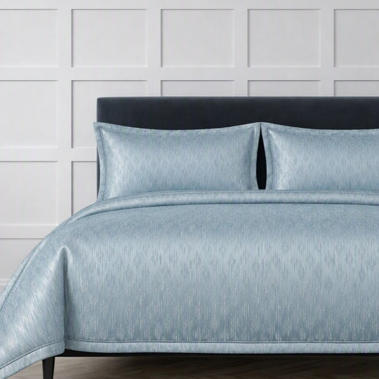 The Judith Leiber Couture Embossed Bedding Set by Ann Gish, shown in light aqua blue, featuring an embossed duvet cover and two matching shams displayed on a bed against a white paneled wall.