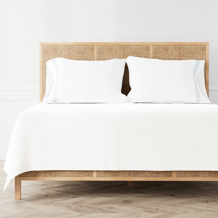 A neatly made bed featuring the Minaudiere Sheet Set in White‑Blue from the Judith Leiber Couture by Ann Gish collection, styled against a light wood rattan headboard and matching footboard in a bright, minimalist room. - SSMIK-WHI-BLE SSMIQ-WHI-BLE