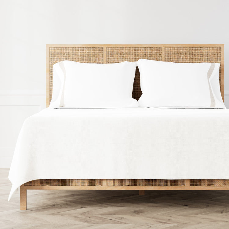 A neatly made bed featuring the Minaudiere Sheet Set in White‑Pumice from the Judith Leiber Couture by Ann Gish collection, styled against a light wood rattan headboard and matching footboard in a bright, minimalist room. - SSMIK-WHI-PUM SSMIQ-WHI-PUM