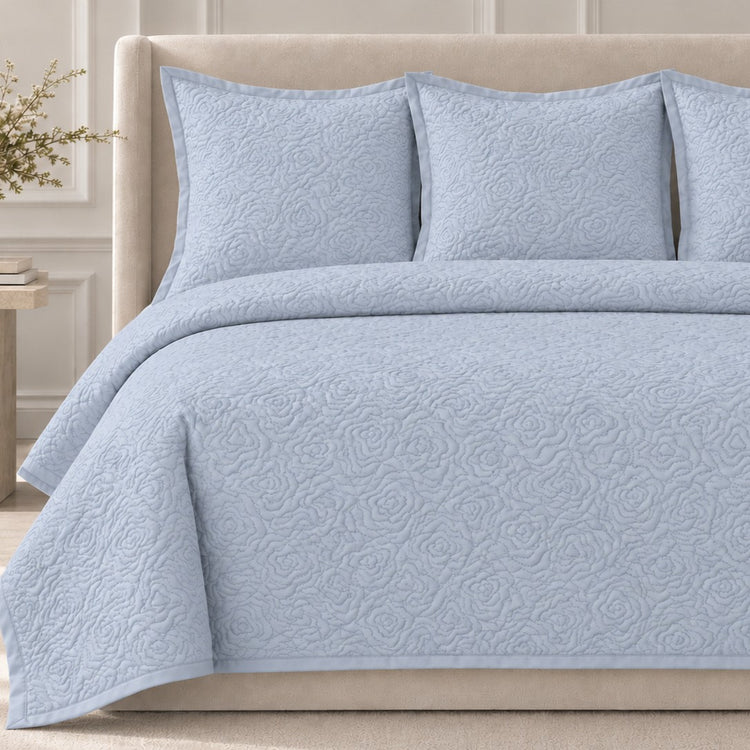 The Roses Coverlet Set from Judith Leiber Couture by Ann Gish, shown in Blue, featuring its signature rose‑quilted pattern with matching shams, styled on a bed with a beige upholstered headboard in a softly lit, paneled bedroom. - YSETCORQK‑BLE YSETCORQQ‑BLE 
