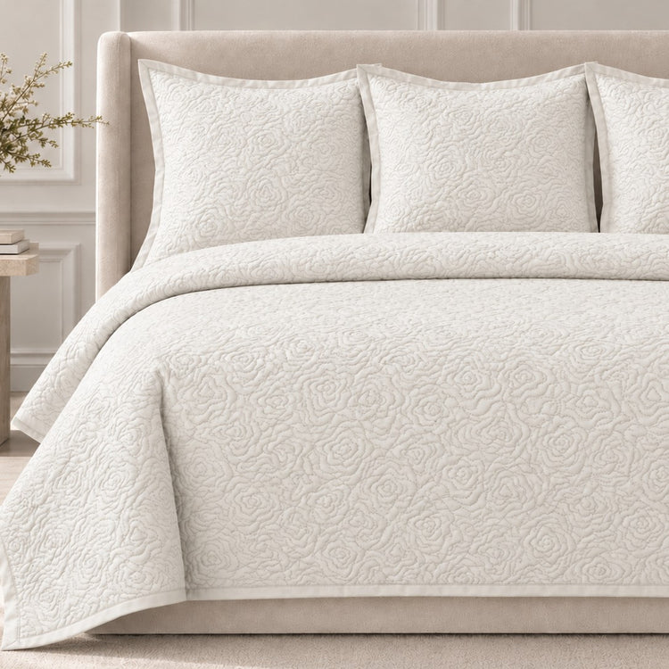 The Roses Coverlet Set from Judith Leiber Couture by Ann Gish, shown in Bone, featuring its signature rose‑quilted pattern with matching shams, styled on a bed with a beige upholstered headboard in a softly lit, paneled bedroom. - YSETCORQK‑BON YSETCORQQ‑BON
