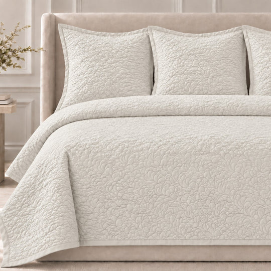 The Shells Coverlet Set from the Judith Leiber Couture by Ann Gish collection, shown in bone with its scalloped quilted pattern, styled on a bed with a beige upholstered headboard and matching shams in a softly lit bedroom. - YSETCOSHK-BON YSETCOSHQ-BON