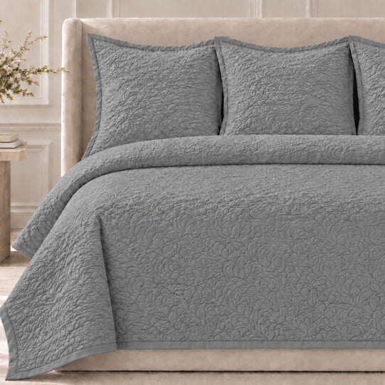 The Shells Coverlet Set from the Judith Leiber Couture by Ann Gish collection, shown in Metal with its scalloped quilted pattern, styled on a bed with a beige upholstered headboard and matching shams in a softly lit bedroom. - YSETCOSHK-MET YSETCOSHQ-MET
